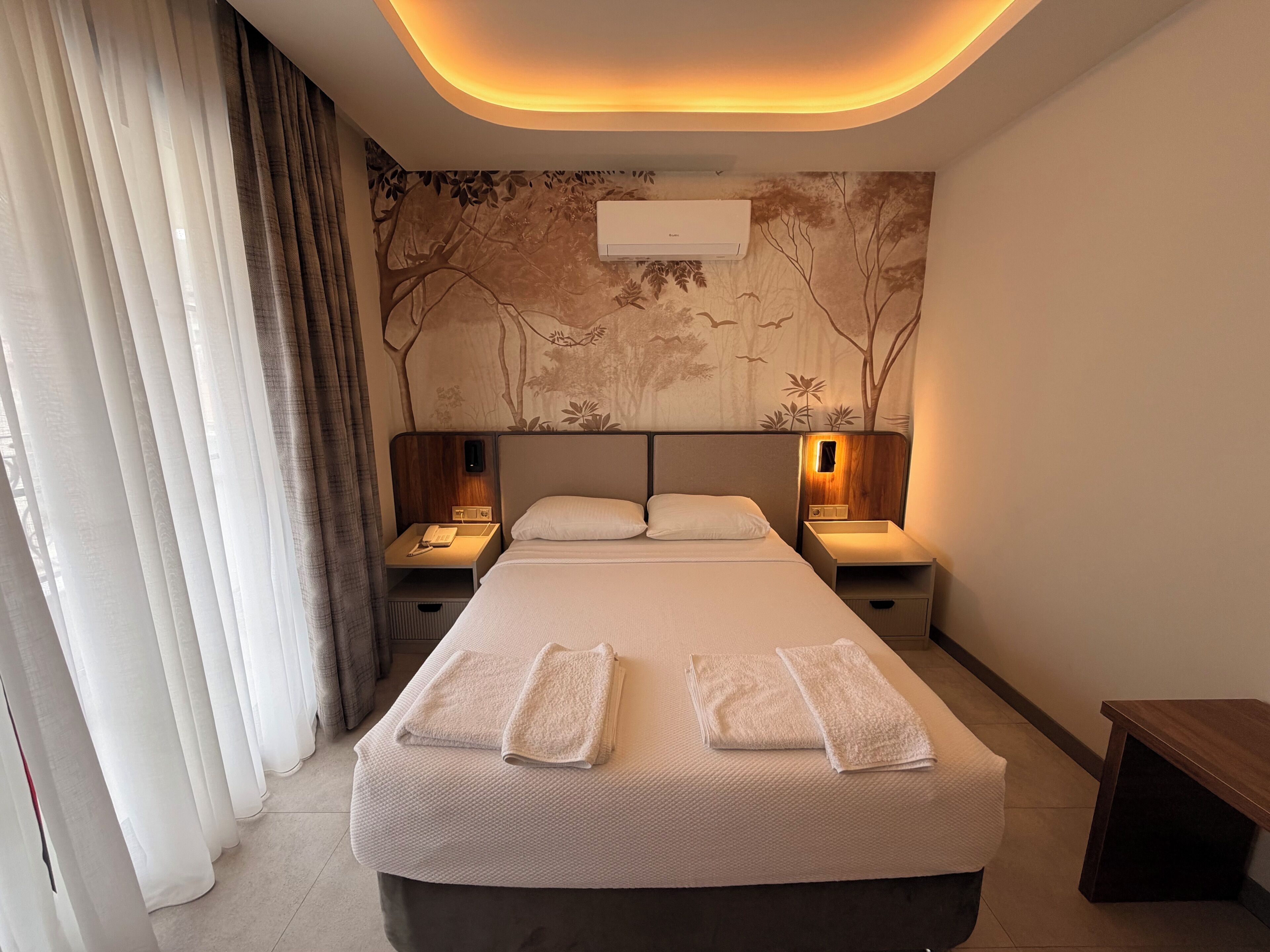 Una habitación de hotel con una cama grande, dos mesitas de noche con lámparas, un aire acondicionado de pared y una cortina en la ventana.