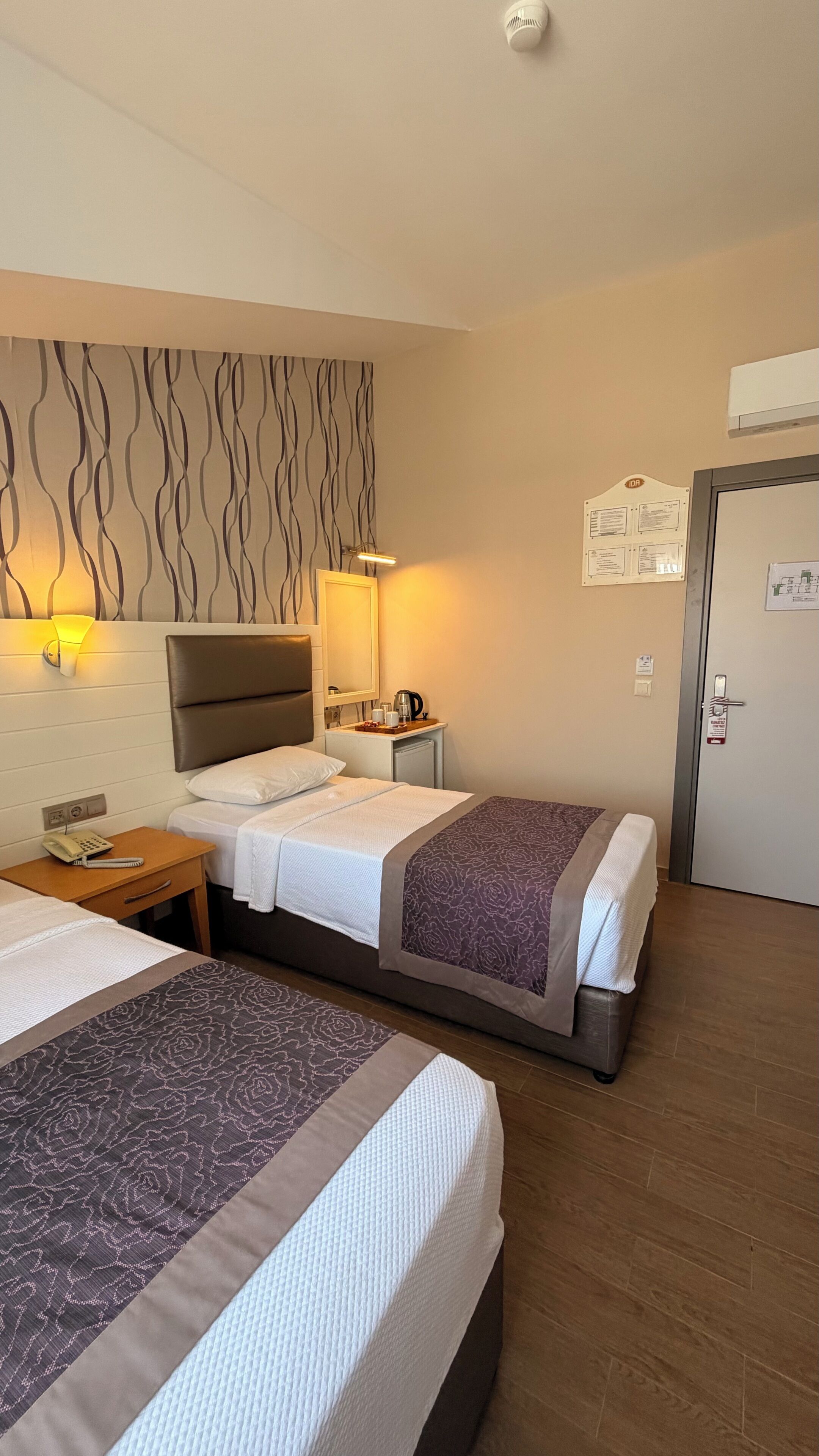 A hotel room with two beds, a bedside table with a lamp, a wall-mounted air conditioner, and a door with a sign.