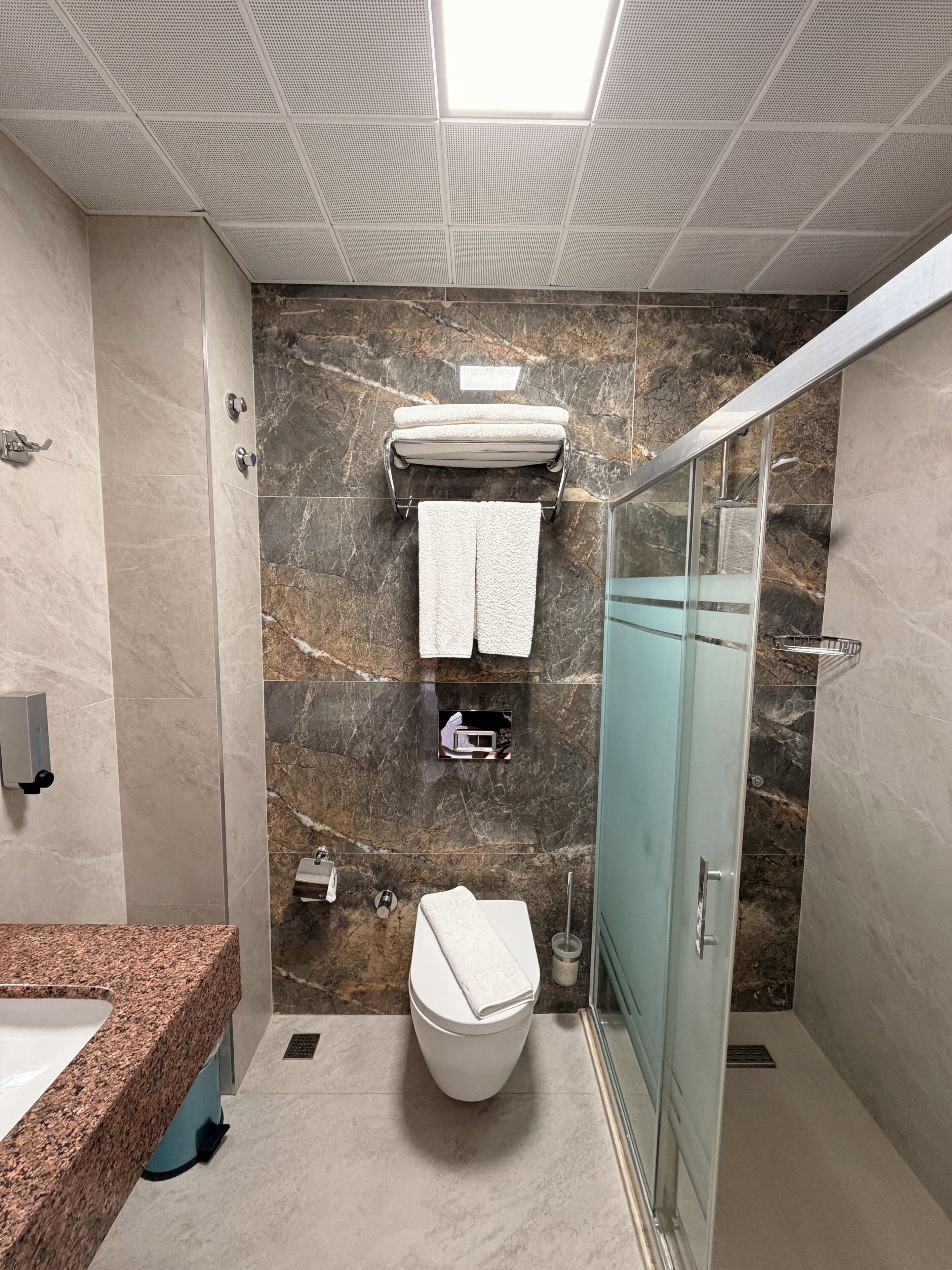 A modern bathroom with a marble wall, a glass shower enclosure, and a granite countertop.