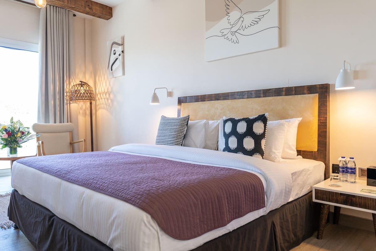 Een opgemaakt bed met een decoratieve plaid, een nachtkastje met een lamp, en een muur met kunstwerken.