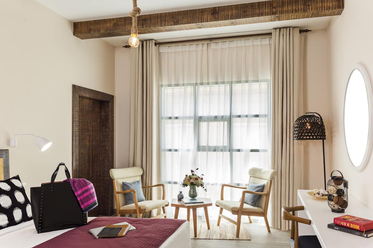 Een hotelkamer met een bed, twee stoelen, een tafeltje, een lamp, een spiegel en een raam met gordijnen.