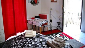 Une pièce comprenant un lit, une table recouverte d’une nappe à pois, une chaise, un rideau rouge, un mur orné d’un tableau représentant des roses et une porte donnant sur l’extérieur.
