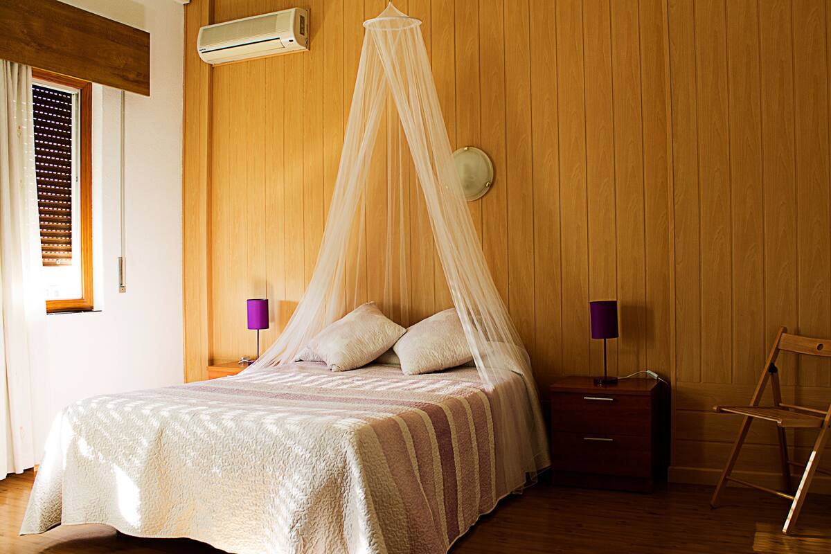 A bedroom with a bed, wooden walls, a nightstand, a chair, and a window with blinds.