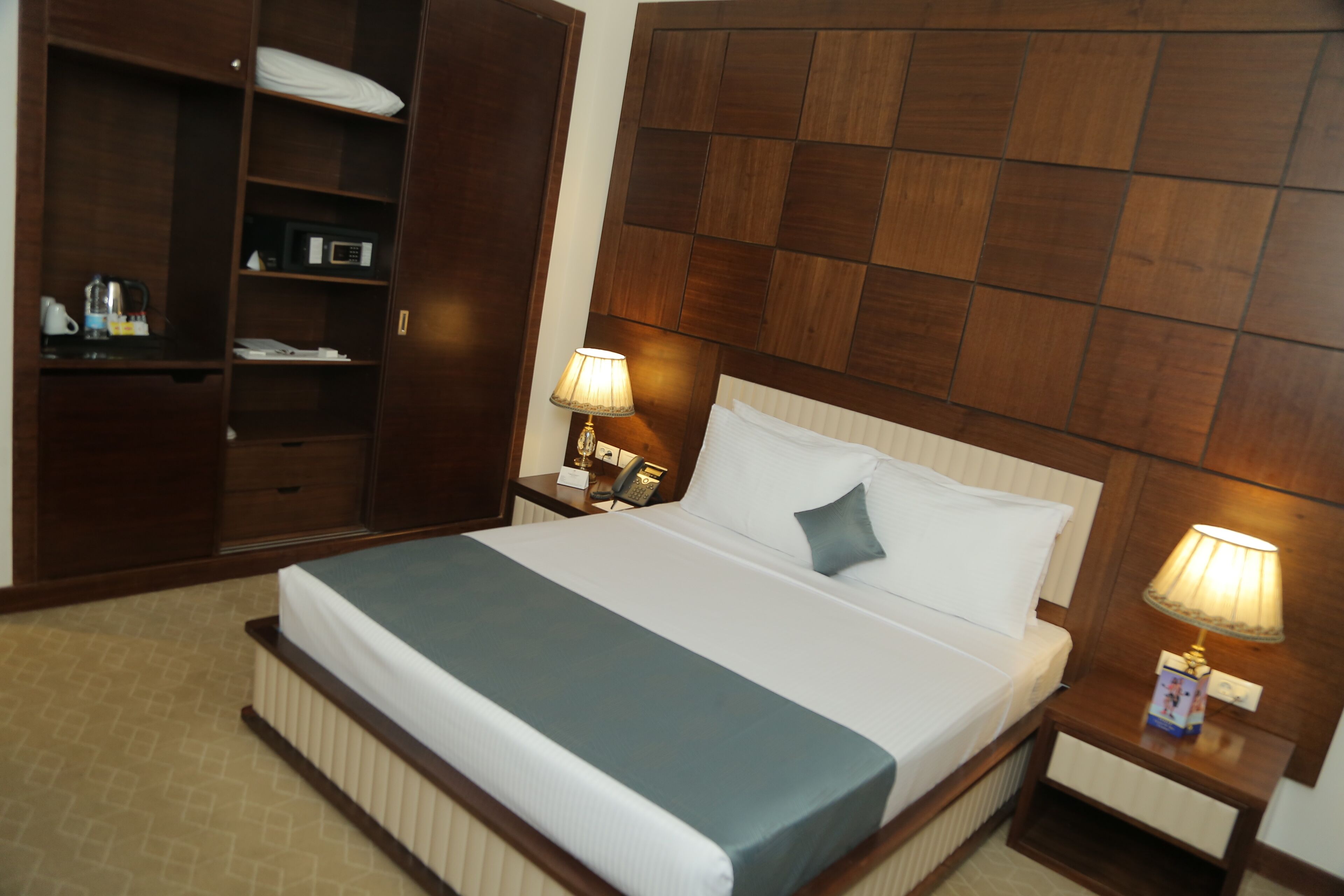 A hotel room with a bed, a nightstand with a lamp, a wardrobe, and a desk with a telephone.