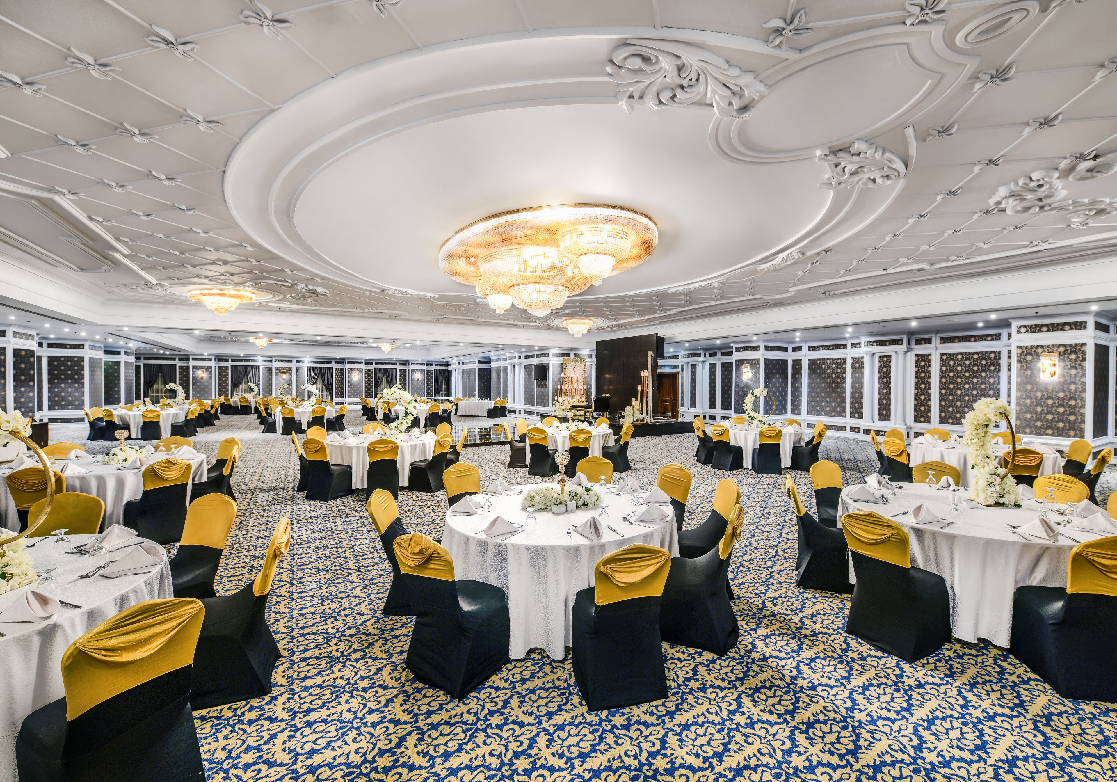 A banquet hall with round tables, black and gold chairs, and a blue patterned carpet.
