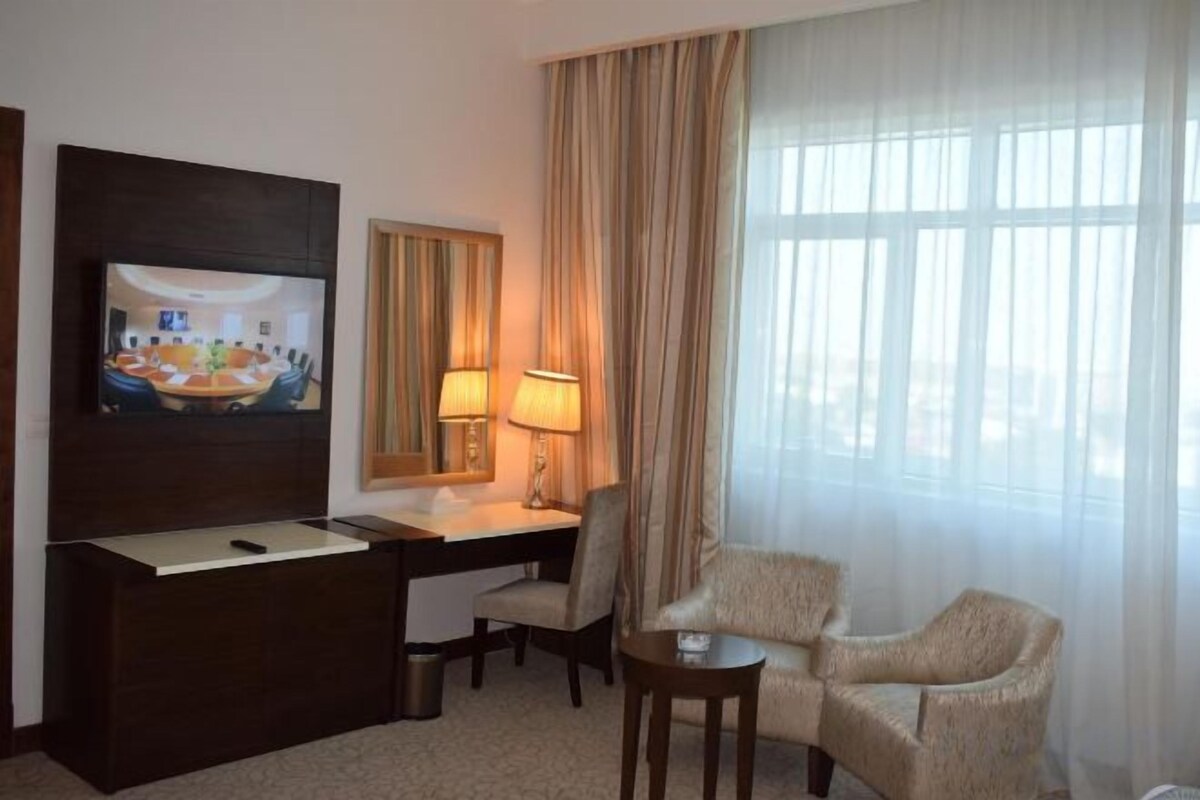 A hotel room with a TV, desk, chair, lamp, and a large window with curtains.