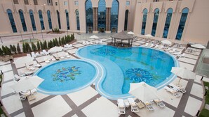 Una amplia piscina interior con una fuente, rodeada de sillas de descanso y sombrillas, y un edificio con grandes ventanales al fondo.