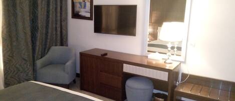 A hotel room with a bed, a desk, a chair, a small stool, a trash can, and a TV mounted on the wall.