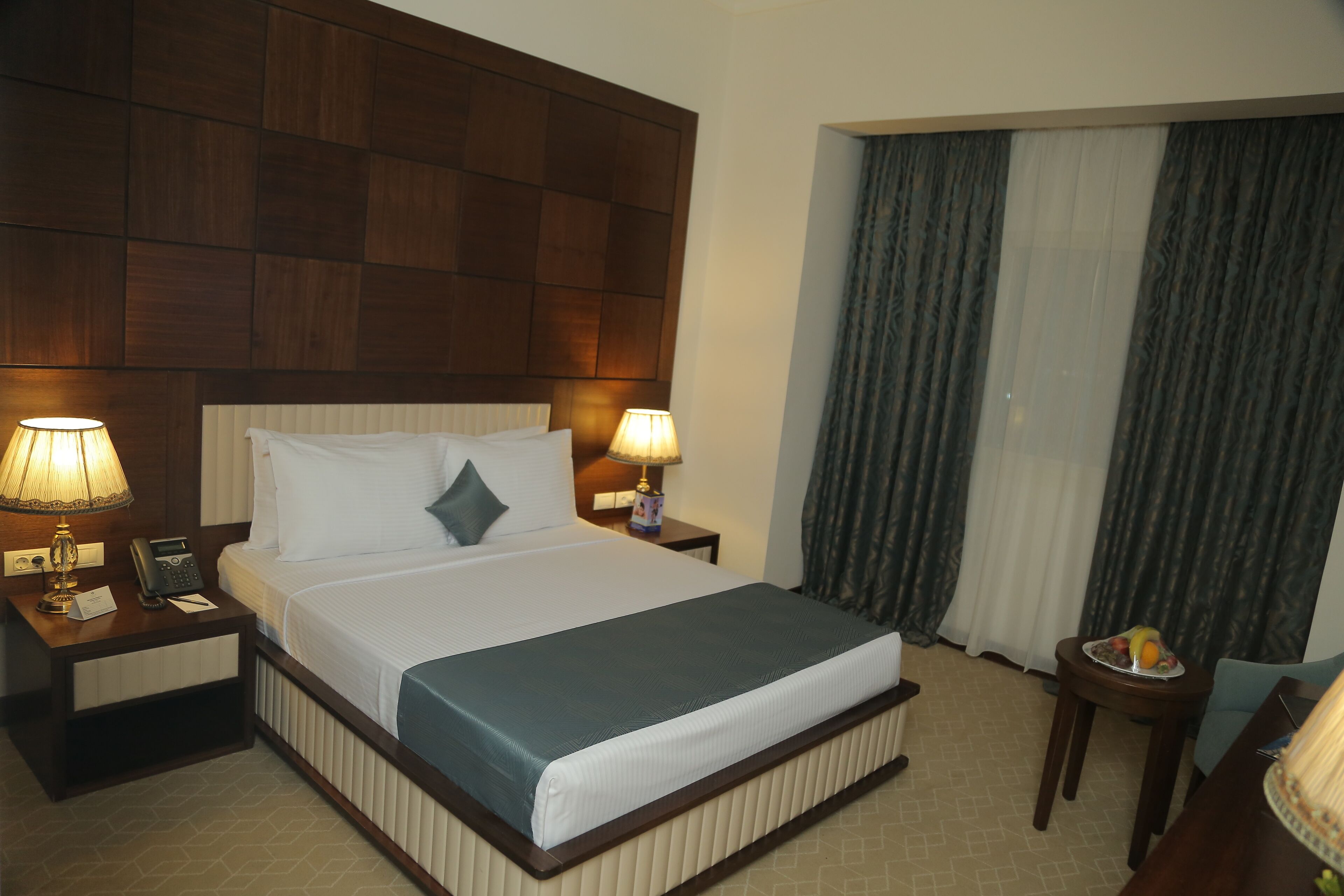 A hotel room with a large bed, two bedside lamps, a telephone, and a small table with a fruit bowl.