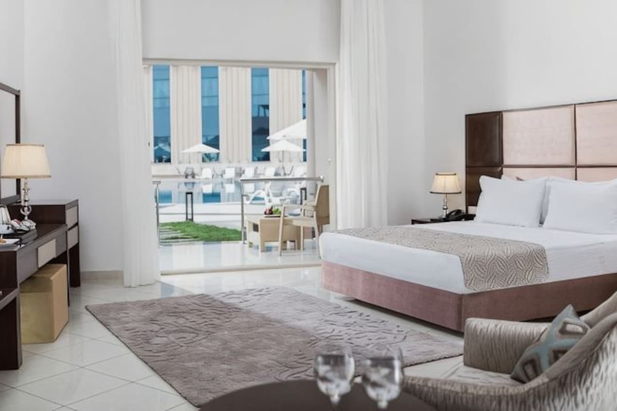 A hotel room with a large bed, a desk with a lamp, a chair, and a view of the outdoors.