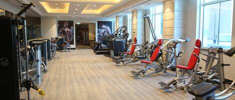 A gym with various exercise machines, a wooden floor, and large windows allowing natural light.