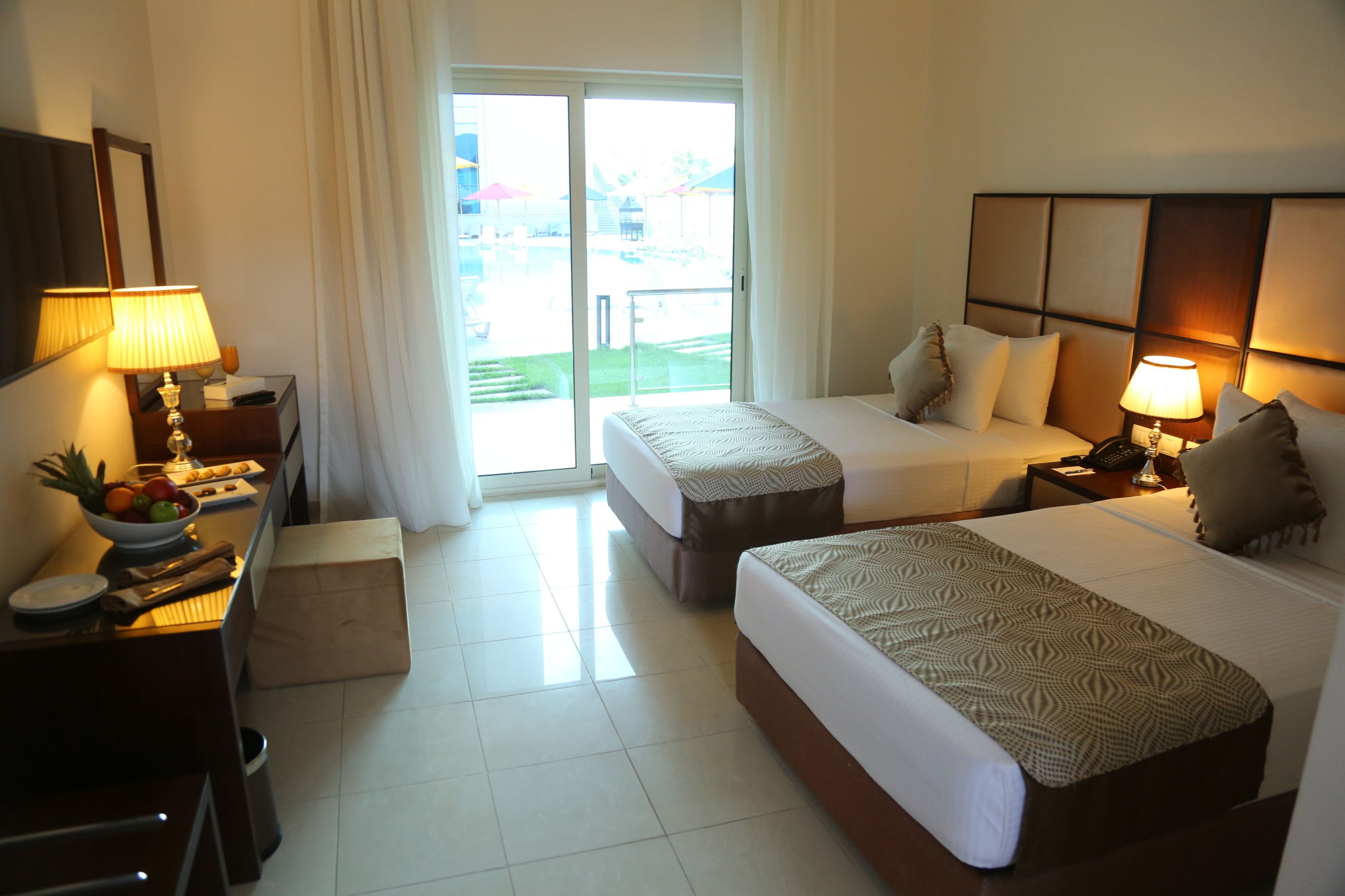 A hotel room with two beds, a desk with a lamp, a small table with fruits, and a view of the outdoors through a sliding door.