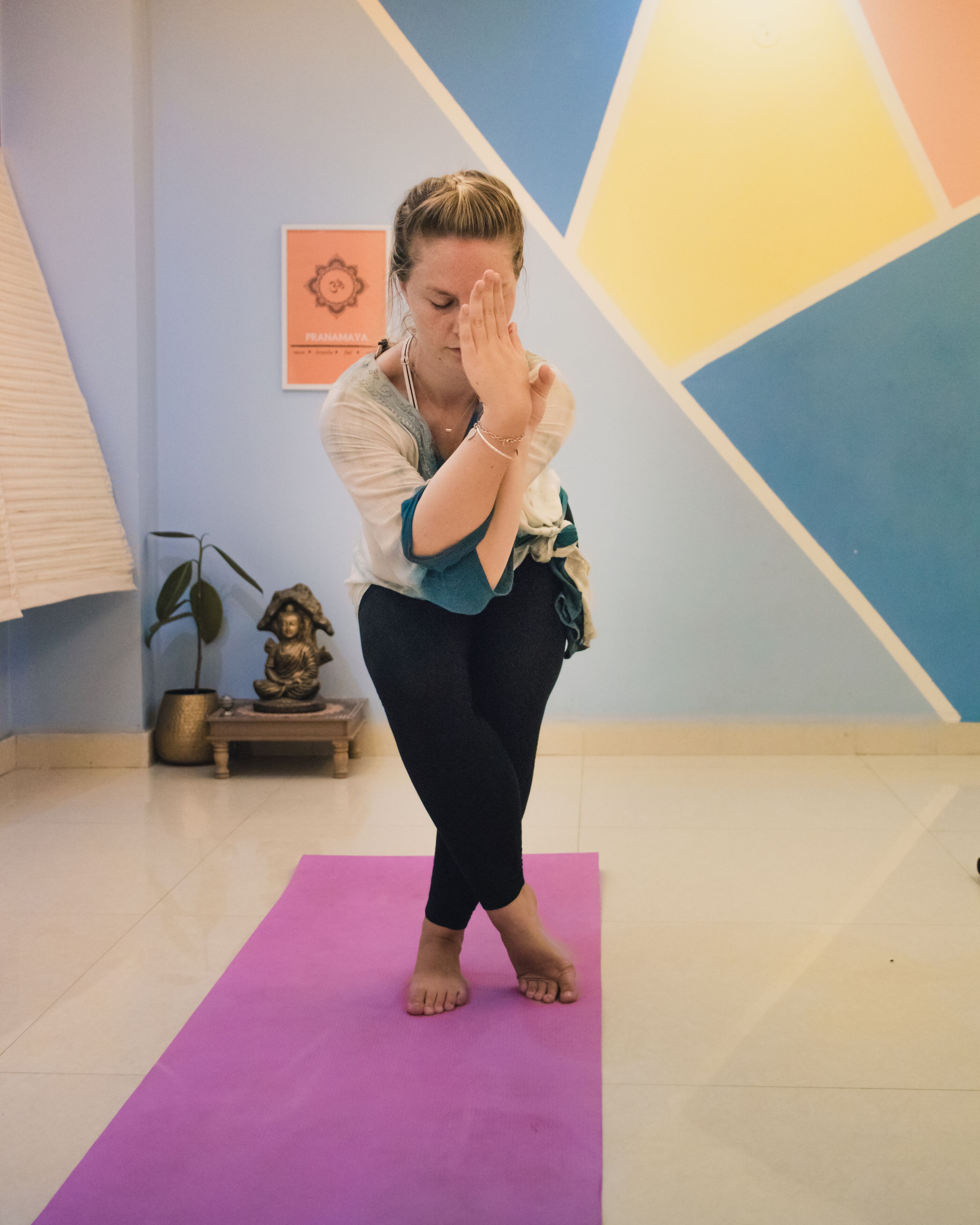 En person utför en yogaposition på en lila matta i ett rum med geometriska mönster på väggen och en liten Buddha-staty.