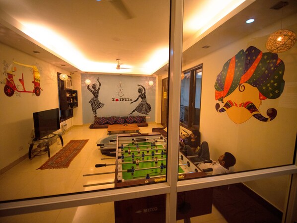 A living room with a sofa, a television, a foosball table, and decorative wall art. - Amigos India - Hostel (New Delhi)