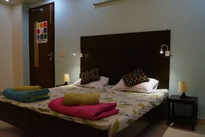 A hotel room with a bed, pillows, a nightstand, and a wall-mounted lamp. - Amigos India - Hostel (New Delhi)