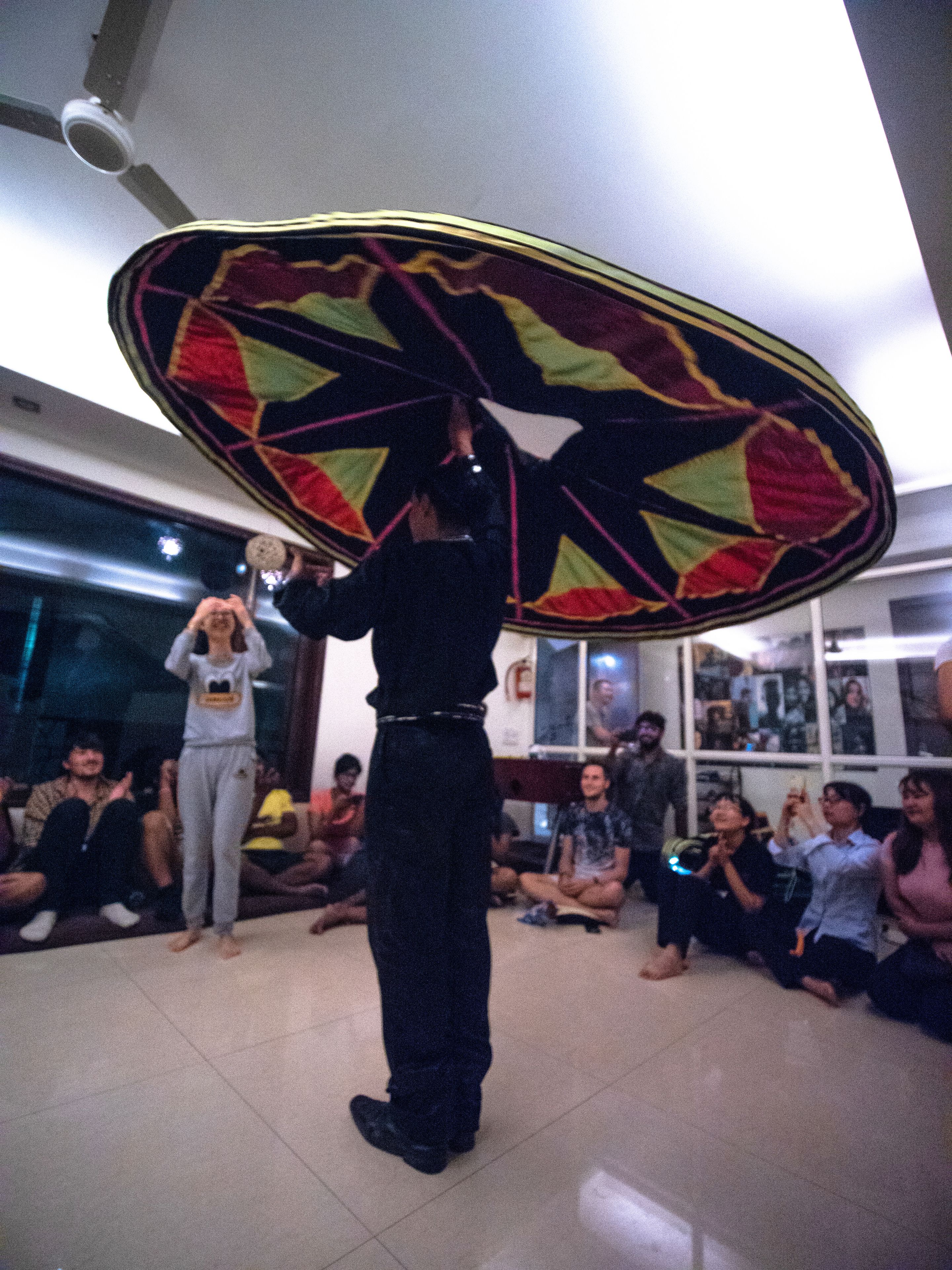 A person performing with a large, colorful sombrero in a room with a tiled floor and a group of seated people.