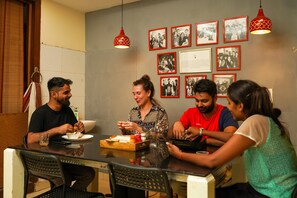 A group of people dining at a table with a bowl of food, a tray, and a bottle of sauce. There are framed pictures on the wall behind them. - Amigos India - Hostel (New Delhi)