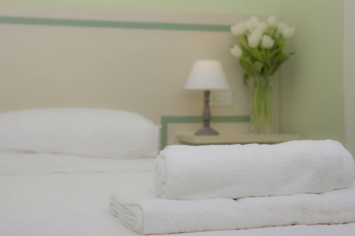A neatly made bed with white towels and a white pillow. There is a nightstand with a lamp and a vase of flowers in the background.