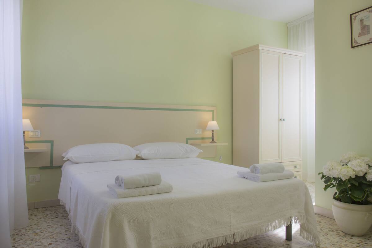 A neatly made bed with white linens and towels, a bedside lamp, a wardrobe, and a potted plant.
