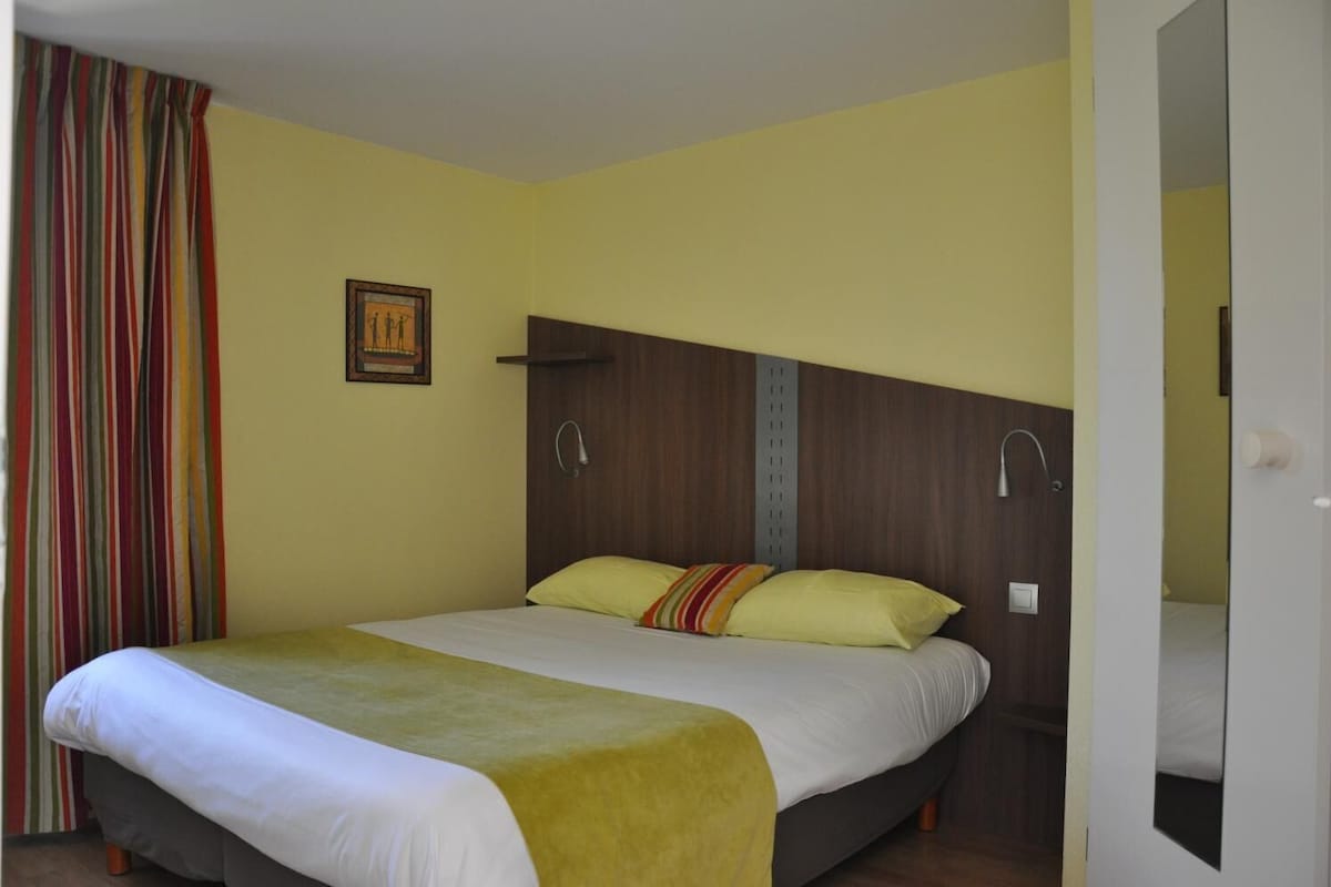 A single-bed room with a wooden headboard, a yellow wall, a striped curtain, and a framed picture.