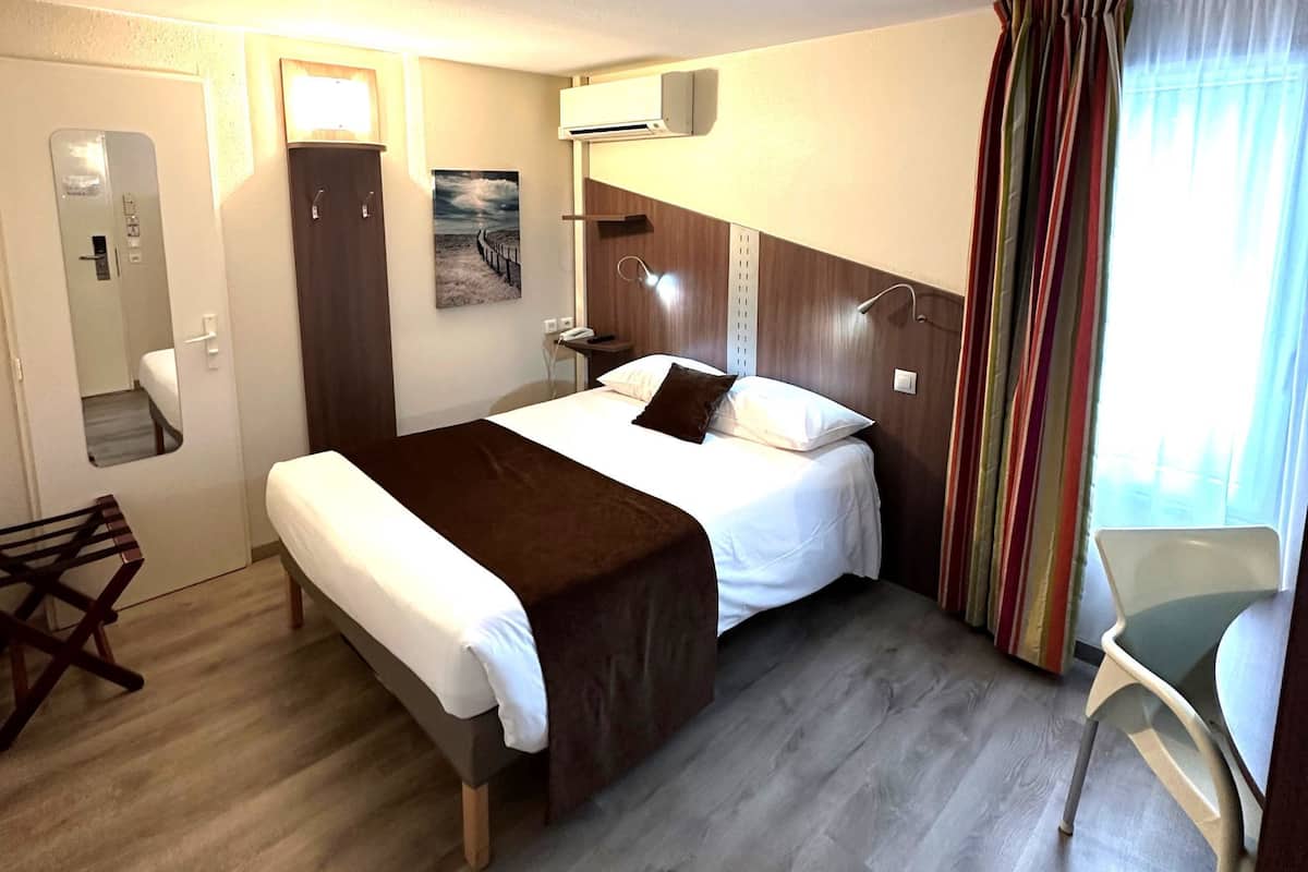 Habitación de hotel con cama, mesita de noche, lámpara y vistas al exterior a través de cortinas.
