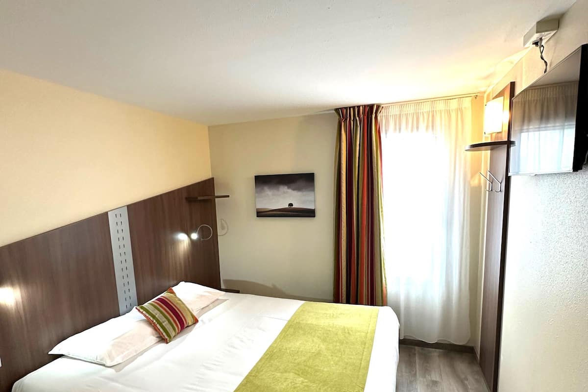 Una habitación de hotel con cama, cabecera, televisor de pared, un cuadro y ventana con cortinas.
