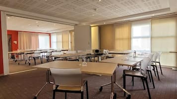 Een vergaderzaal met meerdere tafels en stoelen, een kamerbreed tapijt met een patroon en grote ramen met gordijnen.