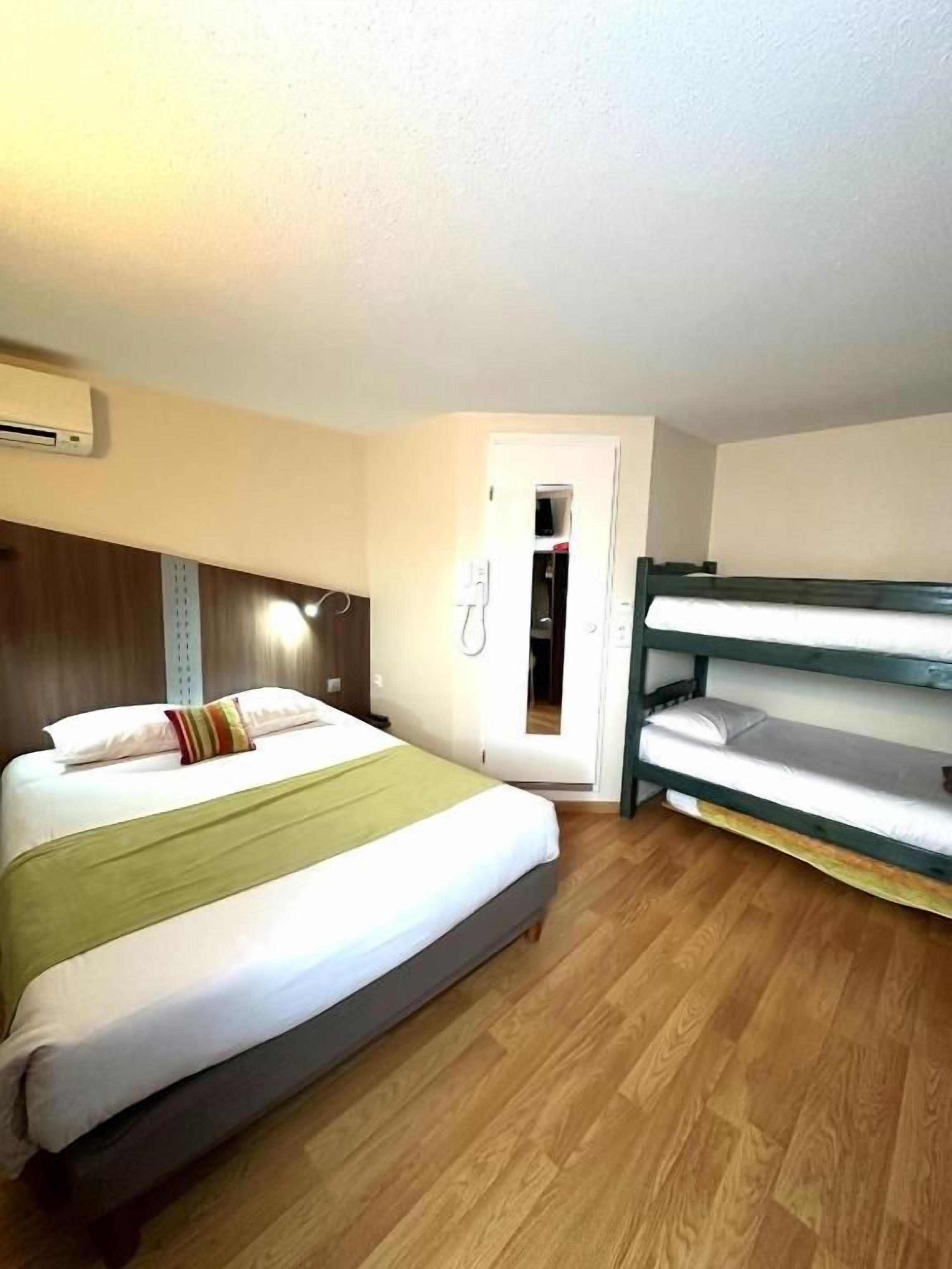 A room with a bed, a bunk bed, wooden flooring, a wall-mounted air conditioner, and a door leading to another room.