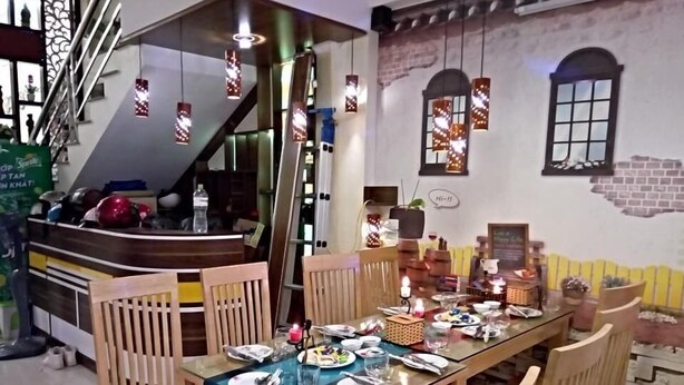 Un espace repas avec une longue table dressée pour plusieurs personnes, des chaises en bois, des lampes suspendues, un escalier et un mur de briques avec une fenêtre.