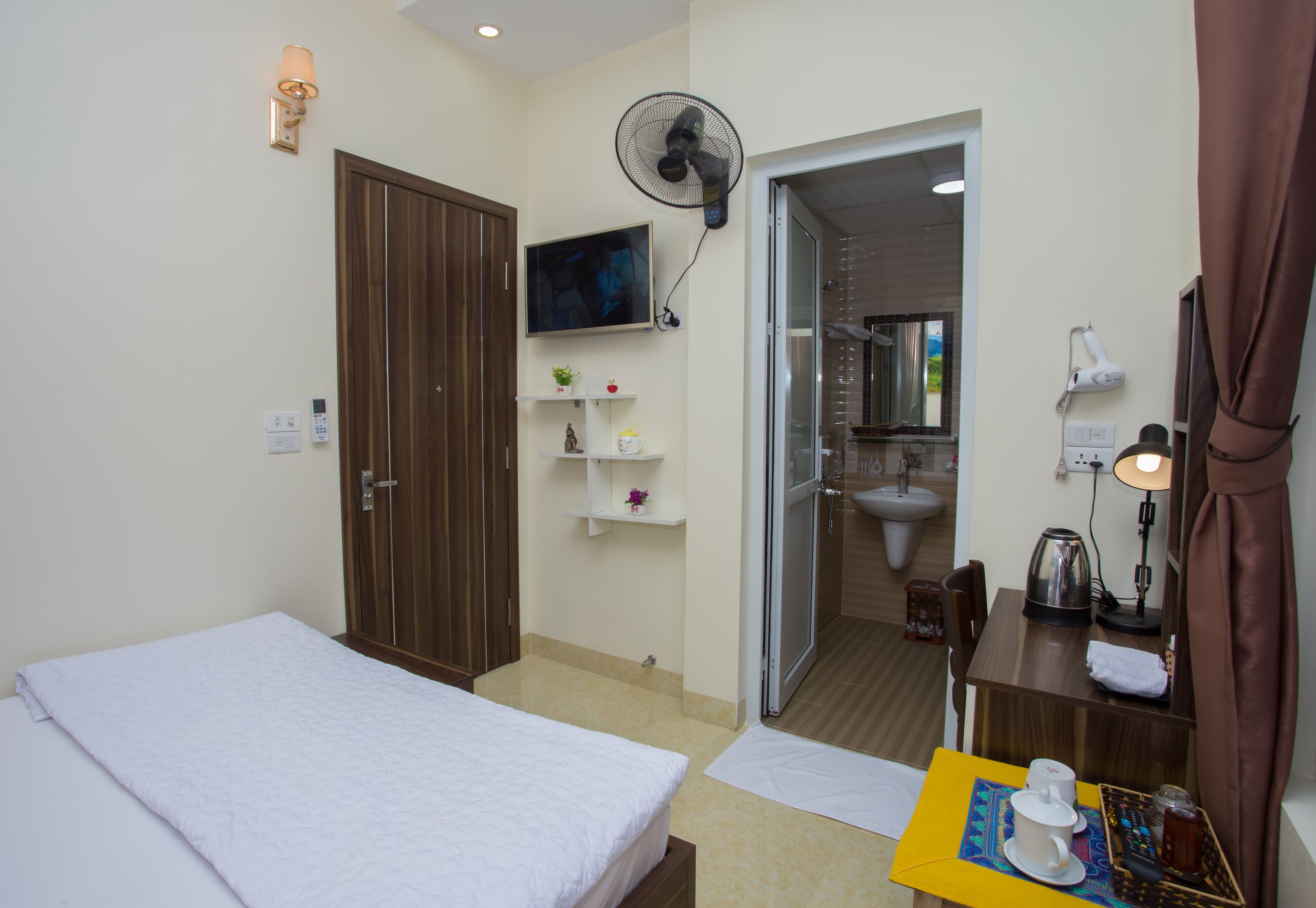 A hotel room with a bed, a desk with a lamp, a fan, a TV, and a bathroom visible through an open door.