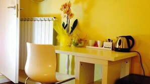 A small, bright room with a yellow wall, a white chair, a small table with a kettle, a mirror, and a TV. - Ager Vaticanus (Rome)