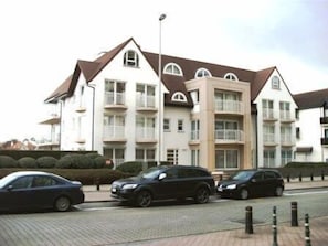 Un edificio residencial de varios pisos con fachada simétrica, balcones y un camino pavimentado con coches estacionados.