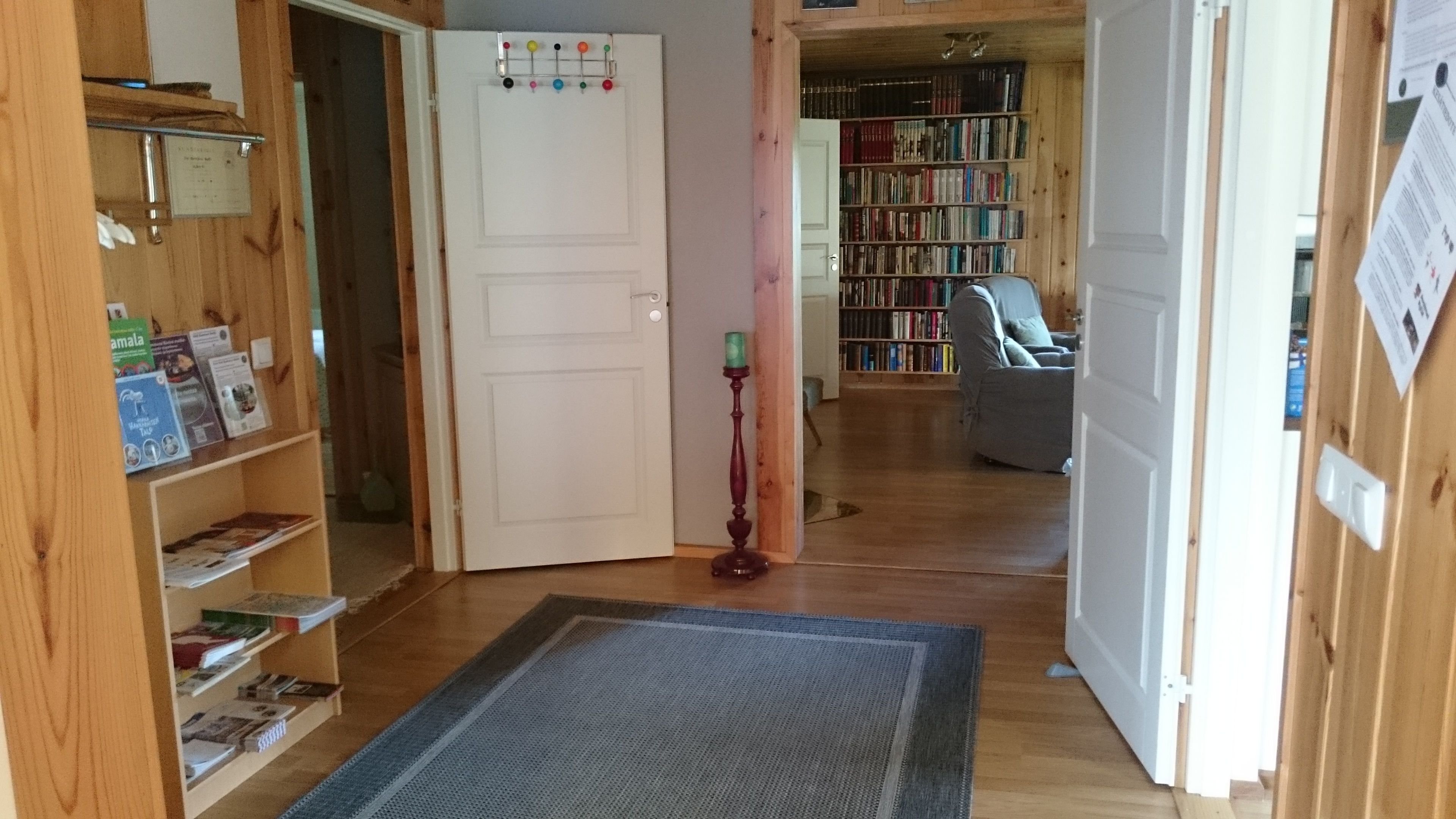 Un couloir au sol en bois, avec une porte, une bibliothèque, un lampadaire et une chaise à l’arrière-plan.