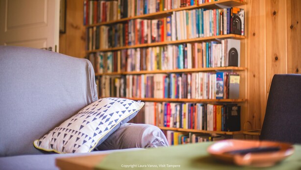 Un rincón acogedor para leer, con un sofá gris, un cojín estampado y una estantería llena de libros.