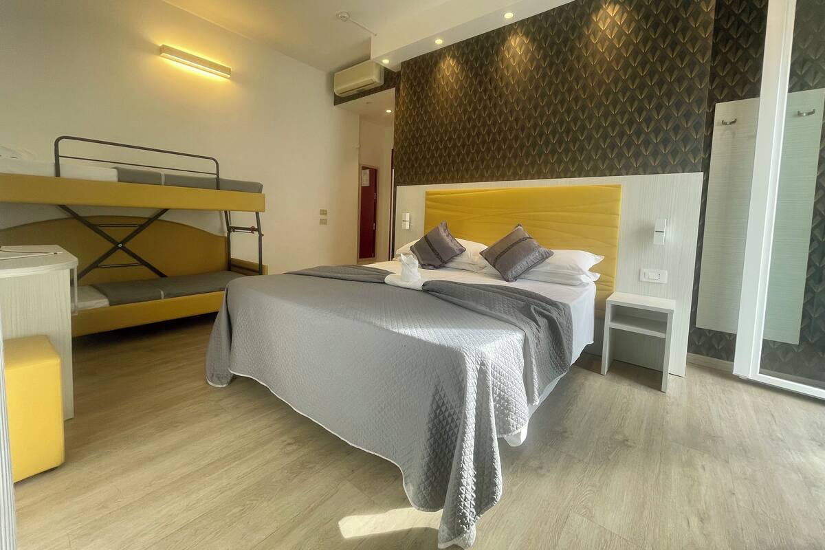 A modern hotel room with a bunk bed, a single bed with a headboard, bedside tables, and a wardrobe.