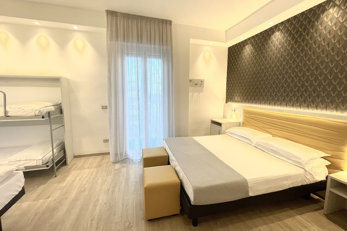 A hotel room with a bunk bed, a single bed, a bedside table, a lamp, and a large window with curtains.