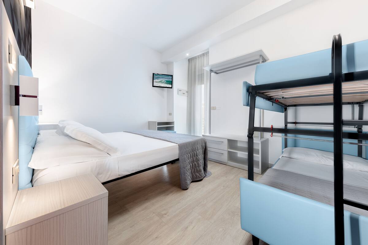 A modern hotel room with a bunk bed, a single bed, a bedside table, a nightstand, a window with curtains, and a wall-mounted air conditioning unit.
