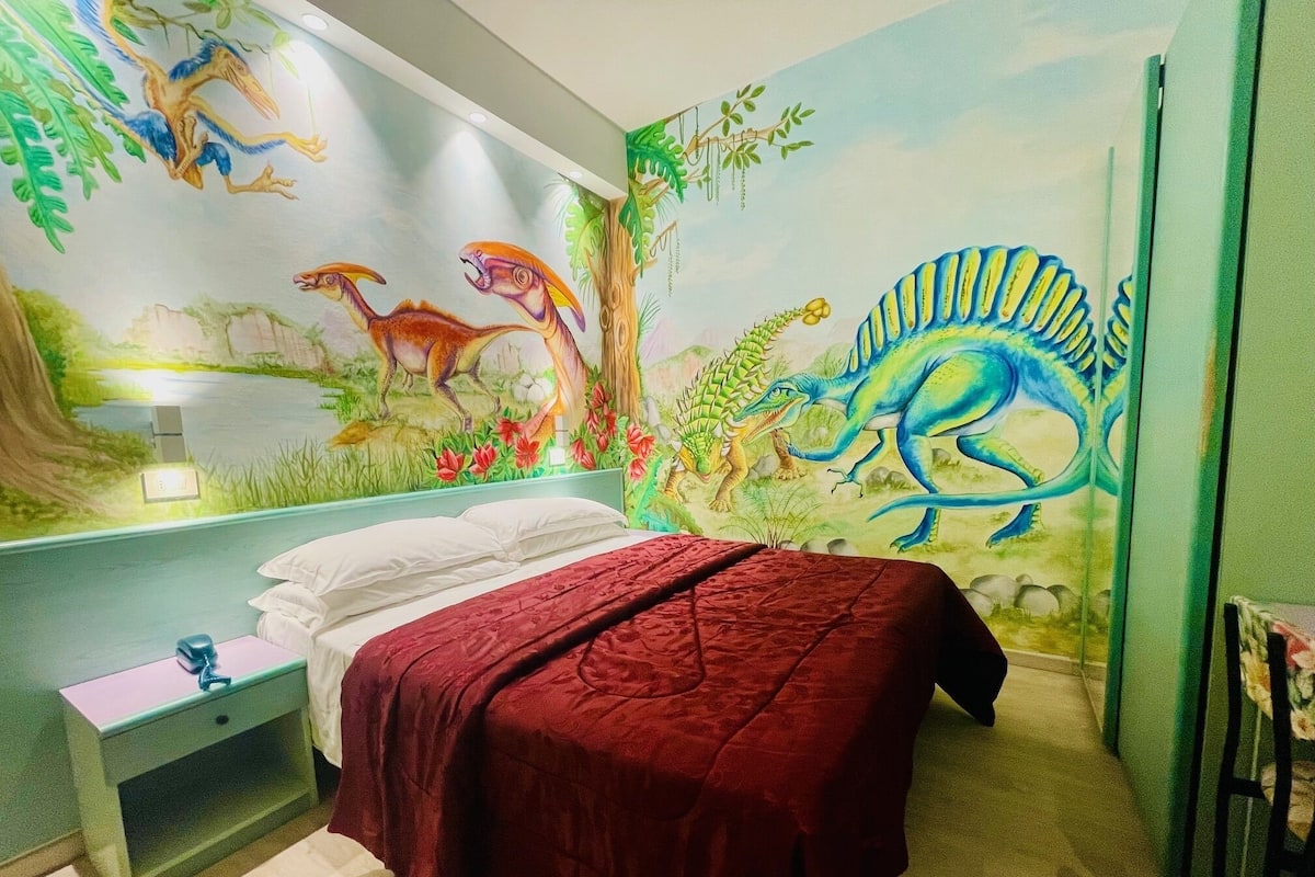 A child's bedroom with a mural of dinosaurs and a bed with a red blanket.