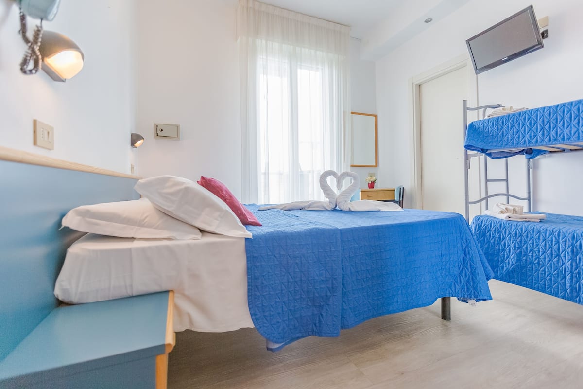 A hotel room with two beds, one with a blue bedspread and the other with a white bedspread. There is a bunk bed with a blue bedspread, a desk with a lamp, and a television mounted on the wall.