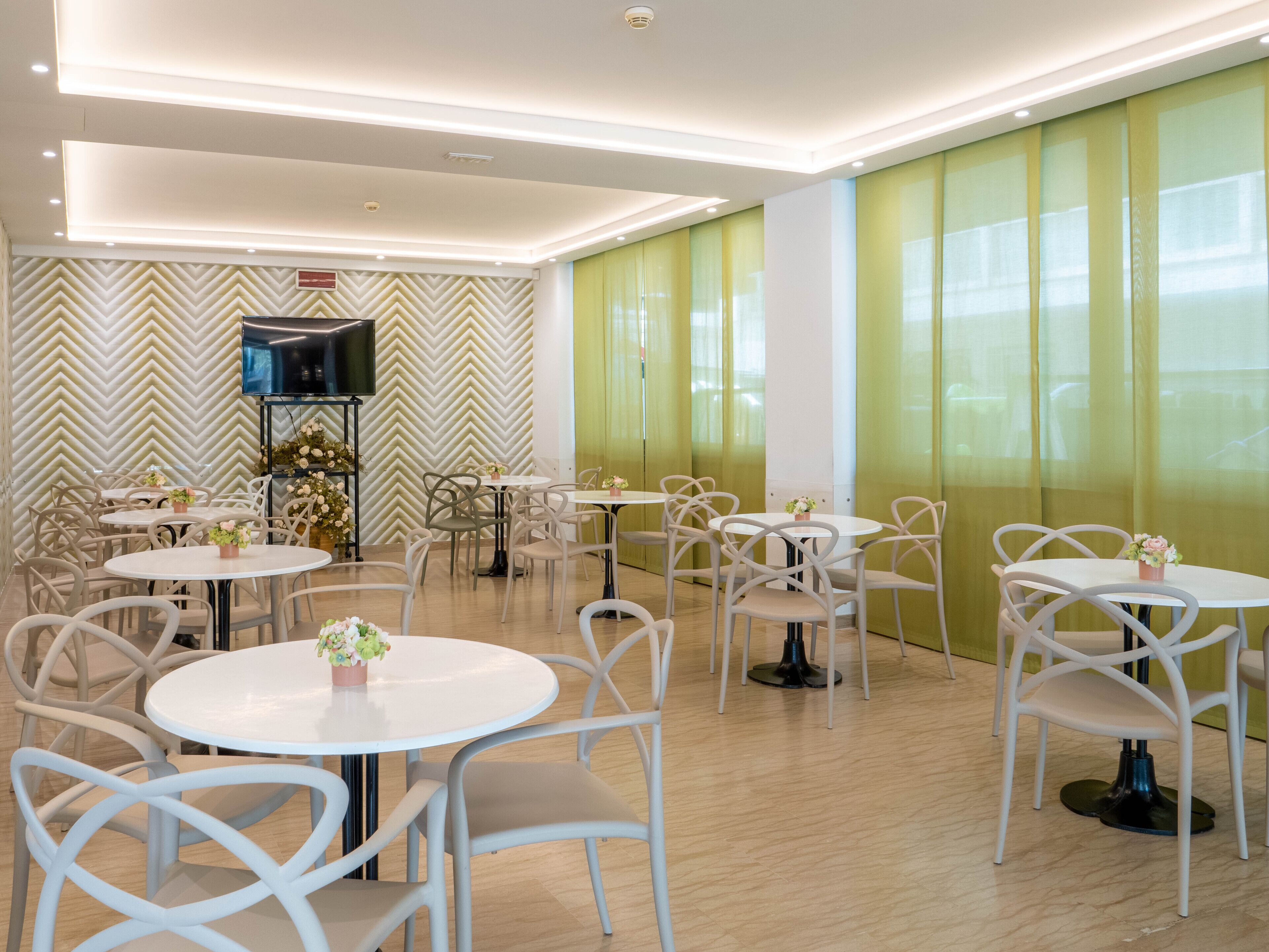 A modern dining area with round tables and chairs, a flat-screen TV mounted on the wall, and a green curtain backdrop.