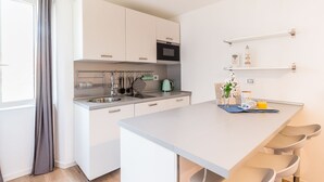 Une cuisine moderne avec des meubles blancs, un évier en acier inoxydable et un four à micro-ondes. Il y a deux chaises autour de la table à manger, un vase avec des fleurs et une petite étagère contenant des ustensiles de cuisine.
