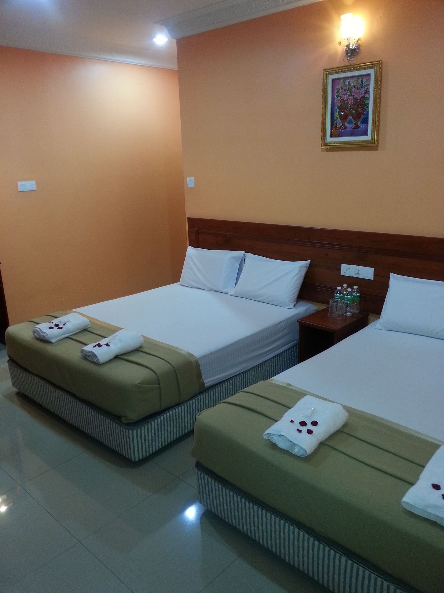 A hotel room with two beds, each with a pillow and a towel. There is a nightstand with a lamp and a small table with bottles.