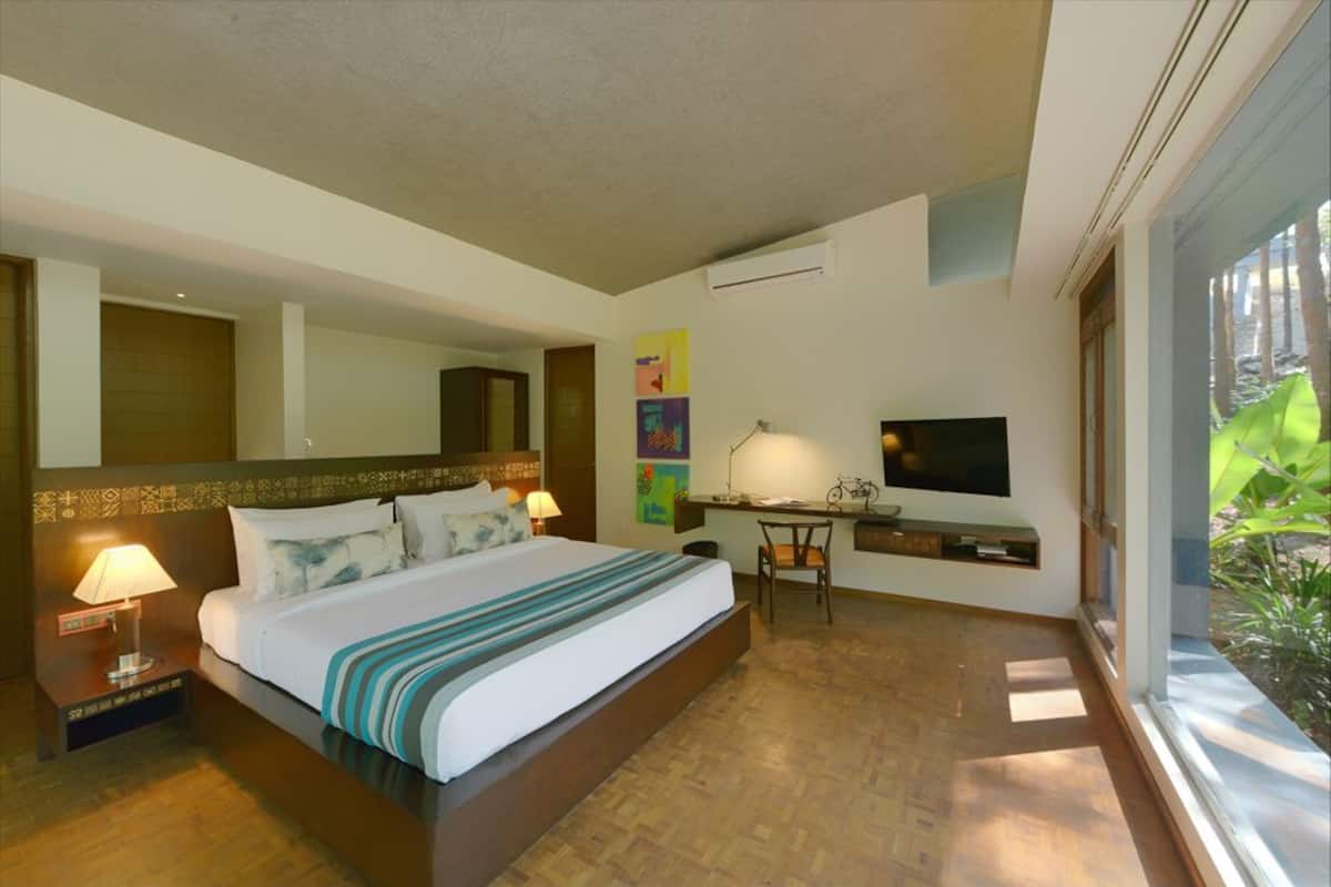 A hotel room with a large bed, a desk, a chair, a TV, and a window with a view of greenery.