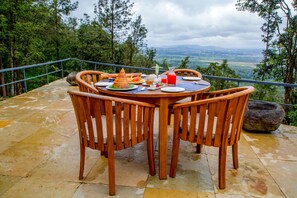 A wooden table with chairs on a stone patio overlooking a forested landscape with a distant view of mountains.