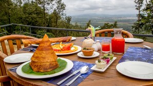 A dining table set with a variety of dishes, including a cone-shaped food item, a bowl of soup, and a glass of red drink, overlooking a scenic landscape. - Java Rain Resorts (Chikkamagaluru)