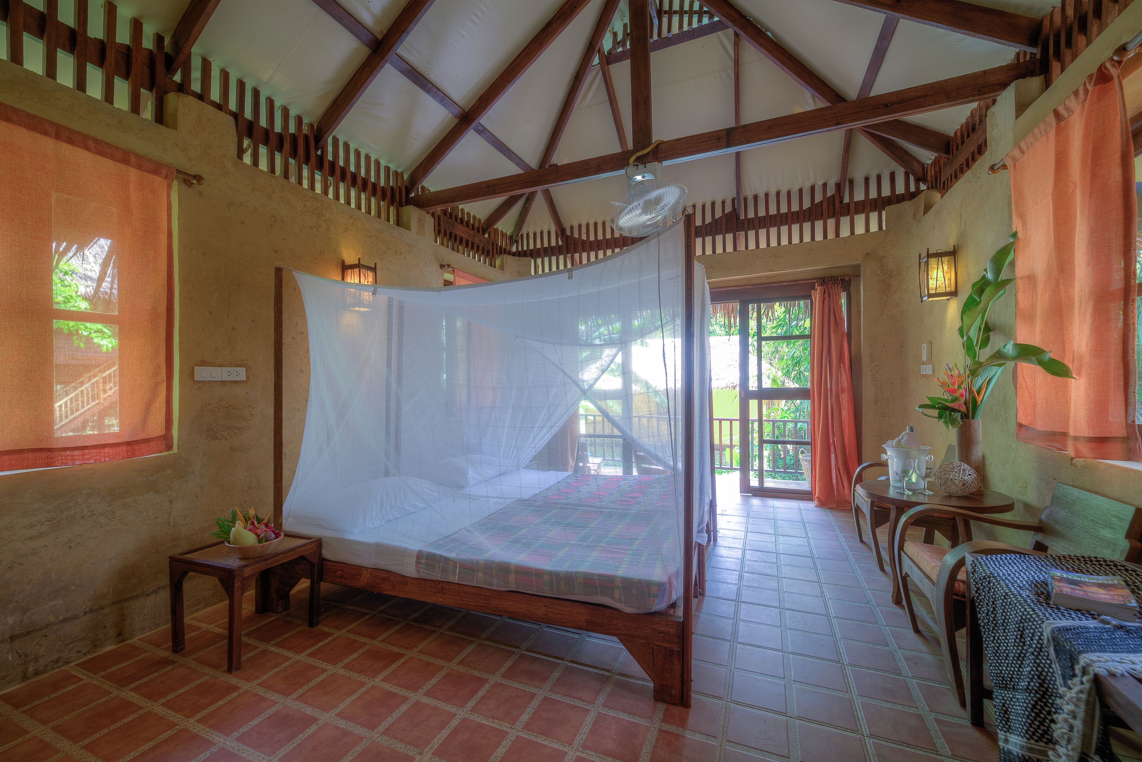 A room with a bed under a mosquito net, a small table with a bowl of fruit, and a bench with a cushion.