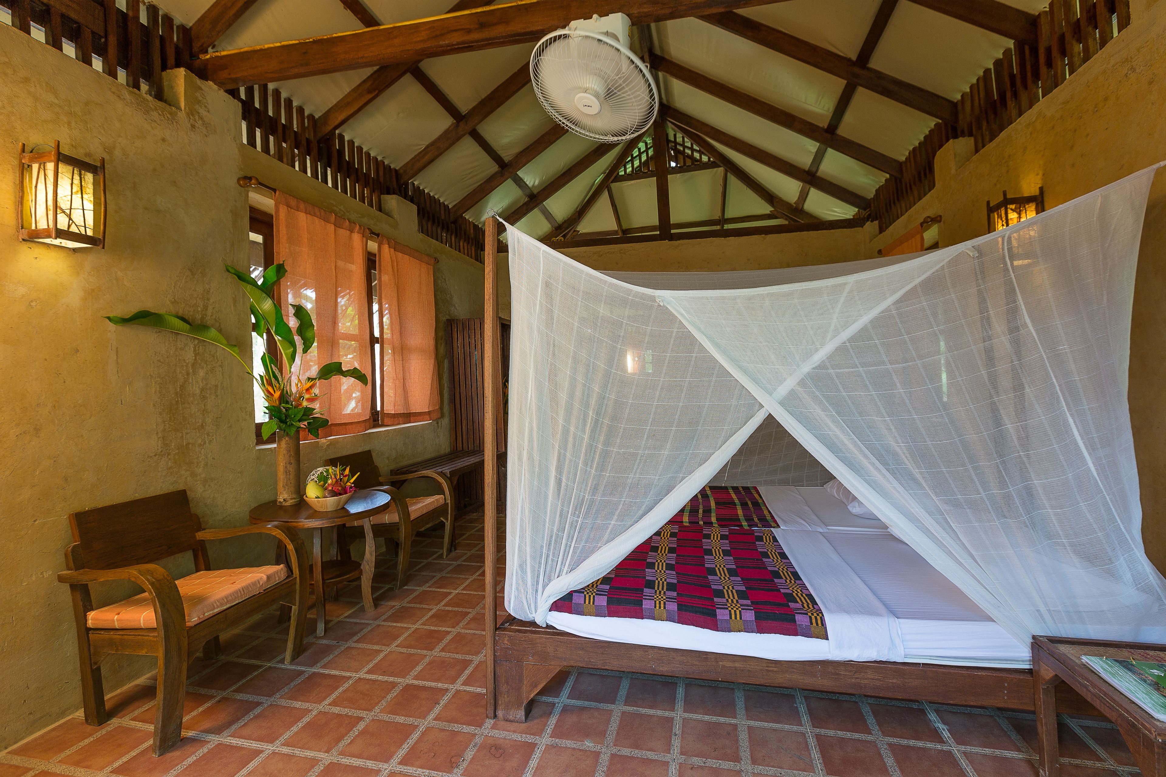 A room with a bed under a mosquito net, a wooden bench, a table with a vase of flowers, and a ceiling fan.
