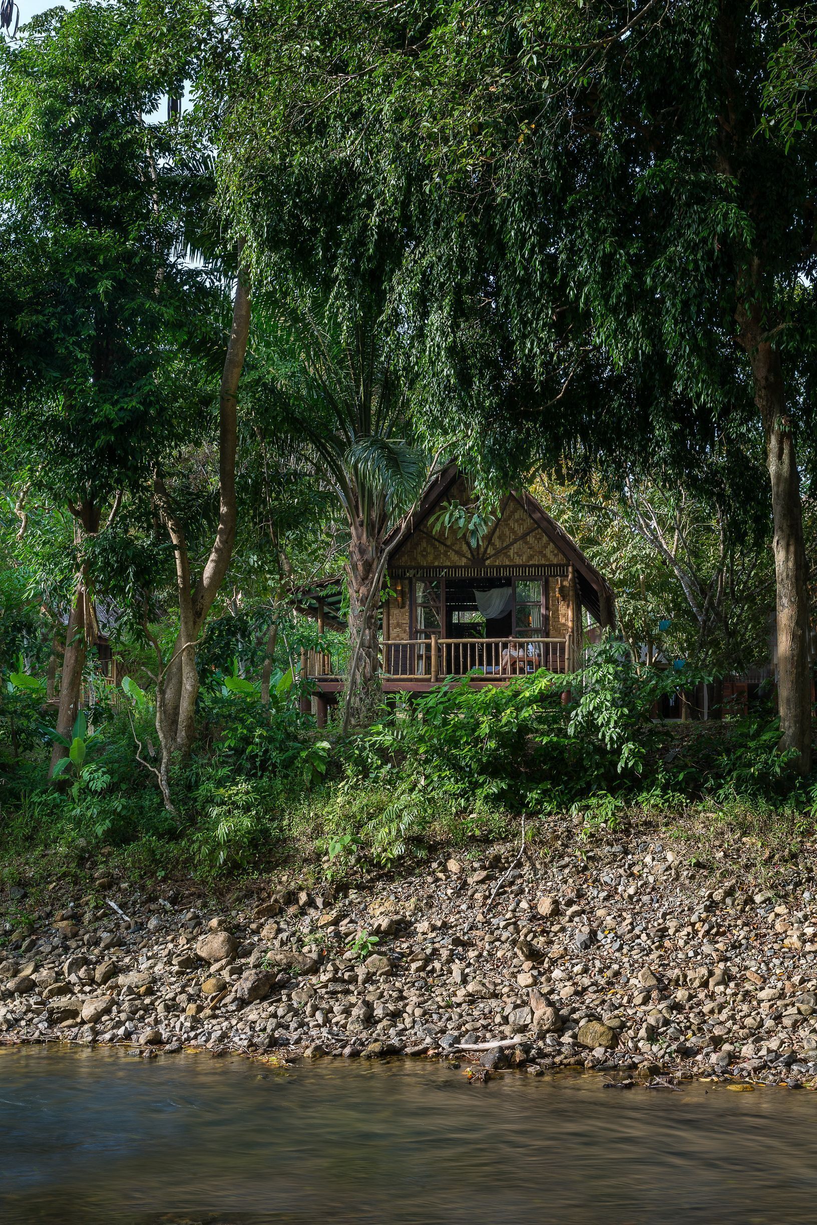 Una cabaña de madera rodeada de un bosque frondoso y la orilla rocosa de un río.