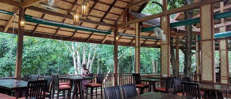 Un área de comedor al aire libre con mesas y sillas de madera, techo de paja y vistas del bosque circundante.