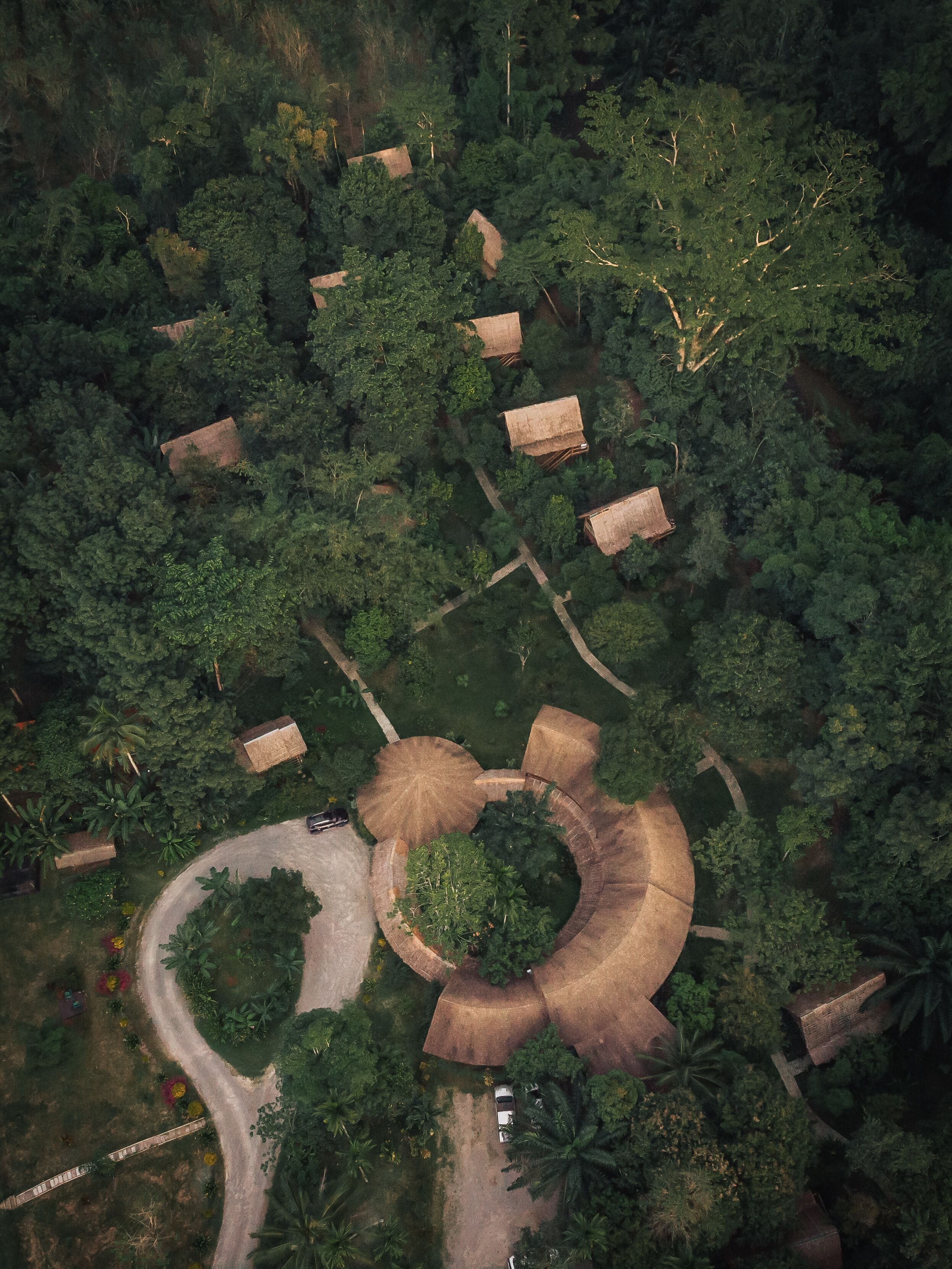 Vista aérea de una zona boscosa con varias cabañas de techo de paja dispuestas en un círculo, rodeadas de abundante vegetación y un sendero serpenteante.